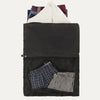 A black Travelpross Essentials washable laundry bag with a large zippered opening, displaying a white shirt and folded plaid and striped garments inside, alongside a mesh pocket with additional items.